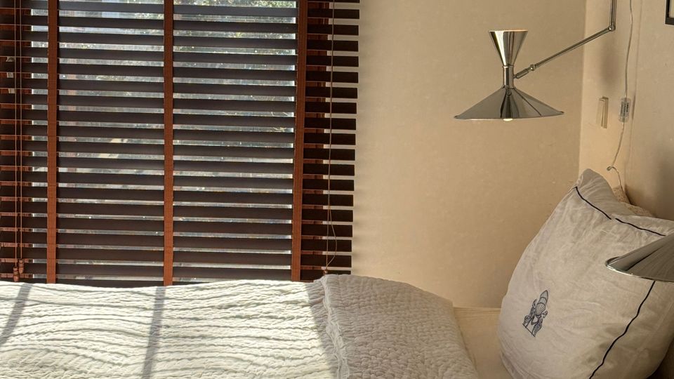 Minimalist bedroom with a neatly made bed in white bedding beside a large window fitted with dark horizontal blinds. Sunlight casts striped shadows across the bed, and a wall-mounted metal reading lamp is positioned above the pillows.