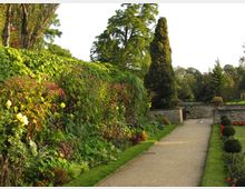 Ein gepflegter Garten mit einem geschwungenen Kiesweg, der durch üppige Blumenbeete und grünes Laub führt. Im Hintergrund befinden sich eine niedrige Steinmauer, eine Brücke und mehrere hohe Bäume.