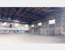 Verlassene Halle mit hohen Ziegelwänden, mehreren eingeschlagenen Fenstern und Stahlträgern im Dachbereich. Die Wände sind mit Graffiti bedeckt, und der Boden ist leer und staubig.