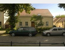 Ein kleines, beigefarbenes Einfamilienhaus mit roten Ziegeldach in einer ruhigen Wohnstraße, flankiert von Bäumen und umgeben von Büschen; vor dem Haus parken zwei Autos auf einer gepflasterten Fläche.