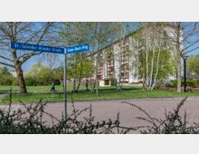 Eine Wohnsiedlung mit einem großen, weißen Mehrfamilienhaus mit roten Fensterrahmen im Hintergrund, umgeben von Bäumen und Grünflächen. Im Vordergrund stehen ein Straßenschild an einer Kreuzung mit den Beschriftungen „Dr.-Salvador-Allende-Straße“ und „Bruno-Baum-Ring“, sowie vereinzelt Spaziergänger auf einem Gehweg.