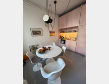 Wohnküche mit pastellrosa Einbauschränken, einem runden weißen Esstisch und vier weißen Stühlen mit gepolsterten Sitzen. An der Wand hängen zwei Bilder, während eine moderne Hängelampe mit schwarzen Kabeln von der Decke hängt. Der Tisch ist mit Obst und Schalen dekoriert.