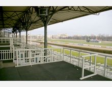 Überdachte Tribüne einer Pferderennbahn mit weißen Geländern und Sitzbereichen. Im Hintergrund sind eine grüne Rennbahn, Werbebanner und mehrstöckige Wohngebäude zu sehen.