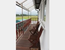 Überdachter Balkon mit Holzboden, zwei klappbaren Holzstühlen und einem Geländer mit roten Ornamenten. Im Hintergrund erstrecken sich grüne Wiesen und ein bewölkter Himmel.