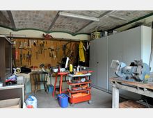 Werkstatt mit gewölbter Ziegeldecke, einer Werkzeugwand mit verschiedenen Hämmern, Schraubenschlüsseln und Zangen, einer roten Rollwerkbank sowie grauen Schränken. Im Vordergrund befindet sich ein Schleifgerät auf einem Holztisch.