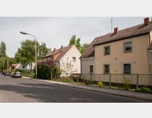 Straßenansicht mit Reihenhäusern aus Putzfassaden und Satteldächern, eingefasst von kleinen Vorgärten mit Zäunen. Im Vordergrund führt eine asphaltierte Straße entlang, geparkt mit vereinzelt stehenden Autos auf der linken Seite, umgeben von Bäumen und Sträuchern.