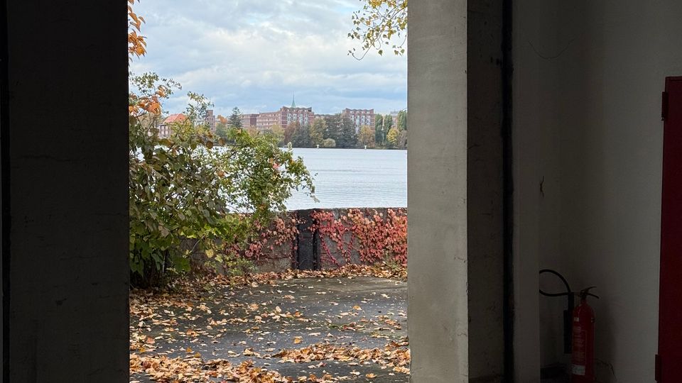 Blick aus einem schlichten, betonierten Innenraum durch eine große rechteckige Öffnung nach draußen auf ein Ufer mit herbstlichem Laub und Sträuchern. Dahinter liegt ein See mit einer gegenüberliegenden Stadt- oder Wohnbebauung unter bewölktem Himmel.