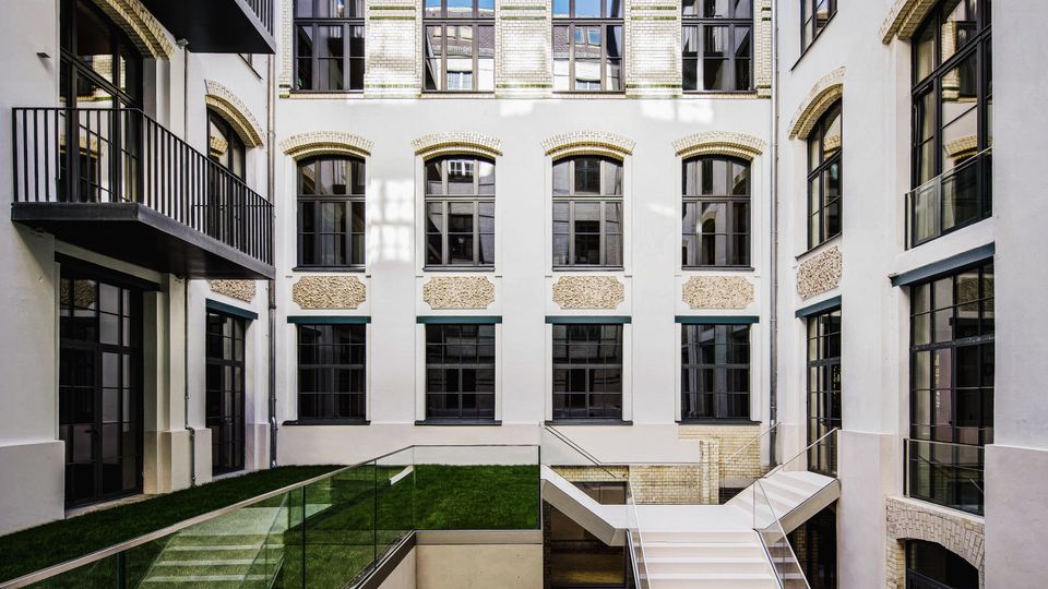 Innenhof eines mehrstöckigen Gebäudekomplexes mit heller Ziegelfassade, großen Sprossenfenstern und seitlichen Balkonen. In der Mitte führen eine breite Treppe und eine Rampe mit Glasgeländern zu einer tieferliegenden Ebene, daneben liegen schmale Rasenflächen.