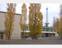 Außenaufnahme des George-C.-Marshall-Hauses, einem modernen Gebäude mit hellbeiger Fassade und großen Glasfenstern, flankiert von hohen Bäumen in Herbstfarben. Im Hintergrund ist der markante Berliner Funkturm zu sehen.