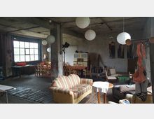 Industrieähnlicher Raum mit groben Betonwänden und sichtbarer Decke, eingerichtet mit verschiedenen Vintage-Möbeln wie Sofas, Stühlen, einem Esstisch und einer Kommode. Große Fenster lassen Tageslicht herein, und Papierlampenschirme hängen von der Decke; die Einrichtung wirkt improvisiert und wohnlich.