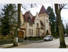 Gelbliches Gebäude im historisierenden Stil mit roten Ziegeldächern, einem hohen Turm, spitzbogigen Fenstern und verzierten Torbögen, umgeben von Bäumen und einer Baumreihe entlang einer gepflasterten Straße mit geparkten Autos.