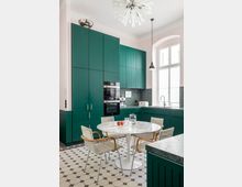 Moderne Küche mit dunkelgrünen Einbauschränken, einem runden weißen Marmortisch und vier Stühlen mit Holzarmlehnen. Der Raum hat ein sechseckiges Bodenfliesenmuster, große Fenster mit viel Tageslicht und eine auffällige Deckenleuchte mit strahlenförmigem Design.