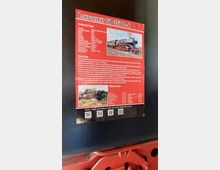 Ein rotes Informationsschild mit der Überschrift „Lokporträt 50 3570-4“, das technische Daten, historische Details und Bilder einer Dampflokomotive enthält. Im unteren Bereich sind QR-Codes für weitere Informationen abgebildet, und der Hintergrund zeigt Teile einer schwarzen und roten Lokomotive.