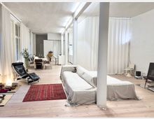 Ein helles, offenes Wohnzimmer mit weiß gestrichenen Wänden und hellen Holzdielen. Im Vordergrund steht ein überzogener Sofa, daneben ein roter Teppich sowie ein schwarzer Liegesessel. Im Hintergrund befinden sich ein Esstisch mit Stühlen, weiße Vorhänge und moderne Dekorationselemente.