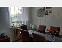 Heller Essbereich mit dunklem Esstisch und vier gepolsterten Stühlen, dekoriert mit Tischläufer, Kerzenhalter und einer Glasvase mit roten Blumen. An der Wand sind weiße Holzpaneele und eine Fotocollage zu sehen; daneben ein Fenster mit transparenten Vorhängen, Fensterbank-Deko und Heizkörper darunter sowie eine runde Hängelampe aus Metallgeflecht.