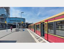 Bahnhof mit einem gelb-roten S-Bahn-Zug auf der rechten Seite und mehreren Bahnsteigen. Im Hintergrund ist ein modernes Gebäude mit Glasfassade und der Schriftzug „Warschauer Straße“ zu sehen, während Passagiere auf dem Bahnsteig unterwegs sind.