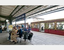 Bahnhof Berlin Frankfurter Allee: Überdachter Bahnsteig mit Sitzgelegenheiten, Abfallbehältern und einer Uhr. Im Hintergrund steht eine geöffnete rote S-Bahn mit ein- und aussteigenden sowie sitzenden Fahrgästen.