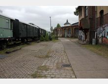 Ansicht eines alten Bahngeländes mit stillgelegten grünen Zugwaggons auf Gleisen links und einem historischen Backsteingebäude mit Rundbögen rechts, teils mit Graffiti besprüht. Der Boden besteht aus Kopfsteinpflaster und Betonwegen, im Hintergrund stehen Bäume und ein spitz zulaufendes Dach.