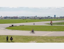 Weite Grünflächen und asphaltierte Wege des ehemaligen Flughafens Tempelhof in Berlin, genutzt von Spaziergängern, Radfahrern und einem Landsegler; im Hintergrund die Skyline der Stadt.