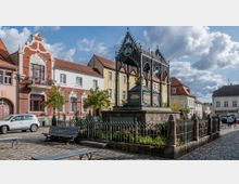 Historischer Marktplatz mit einem kunstvollen gusseisernen Brunnenpavillon auf einem steinernen Sockel, umgeben von einer niedrigen Metallzaunanlage. Im Hintergrund sind restaurierte Altbaufassaden mit roten Ziegeln und hellen Putzflächen sowie gepflasterte Wege und Sitzbänke zu sehen.