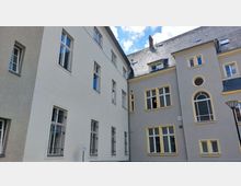 Hofansicht eines mehrstöckigen Gebäudekomplexes mit hellen verputzten Fassaden, großen Fenstern und akzentuierten Rahmungen in Beige und Gelb. Im Hintergrund ist ein graues Schieferdach mit Gauben und ein kleiner Schornstein zu sehen.