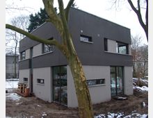 Zweistöckiges, modernes Wohnhaus mit einer Fassade in Dunkelgrau und Beige, großen Fenstern und einem flachen Dach, umgeben von kahlen Bäumen und schmelzendem Schnee auf dem Boden. Im Vordergrund ist ein Baum mit hellem Stamm sichtbar.