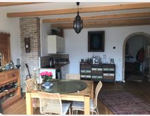 Rustikales Wohnzimmer mit Holzdeckenbalken, Holzboden und gemauerter Backsteinwand. Im Vordergrund steht ein Esstisch aus Holz mit geflochtenen Stühlen, dahinter befinden sich eine Küche mit weißen Schränken und ein Sideboard mit bunten Schubladen. Ein Torbogen führt in einen weiteren Raum.