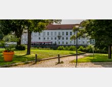 Ein weißes, mehrstöckiges Gebäude mit rotem Dach, beschriftet als "Schloss Restaurant", im Hintergrund einer gepflegten Grünanlage mit gepflastertem Weg, Bäumen und einem großen orangenen Blumentopf. Im Vordergrund sind eine Straßenlaterne und ein Schild zu sehen.