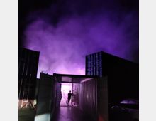 Eine nächtliche Szene zeigt zwei aneinander gereihte Frachtcontainer, zwischen denen ein Gang verläuft. Im Hintergrund steht eine Person im neonvioletten Licht, umgeben von aufsteigendem Nebel.
