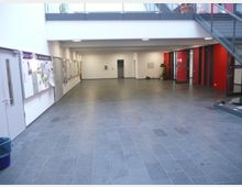 Ein heller Innenbereich eines öffentlichen Gebäudes mit grauem Fliesenboden, weißen Wänden und roten Akzentelementen. An der linken Wand befinden sich Pinnwände mit Informationen, rechts führt eine Treppe nach oben, und im Hintergrund sind Türen und Pflanzen sichtbar.