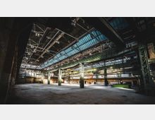 Innenraum einer großen, verlassenen Industriehalle mit hohen Decken aus Stahlträgern und Glasoberlichtern. Das Gebäude zeigt metallene Stützpfeiler, Backsteinwände und eine weitläufige, leere Fläche am Boden.
