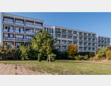 Mehrstöckiges Wohngebäude mit Balkonen und großen Fensterfronten, umgeben von Bäumen, Sträuchern und einer Grünfläche im Vordergrund. Der Himmel im Hintergrund ist klar und blau.