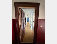 Blick durch einen dunkelbraunen Türrahmen in einen langen, schmalen Flur mit hellem Holzboden und weißen Wänden. Entlang des Gangs befinden sich mehrere Türen, eine Deckenlampe und am Ende eine blau gestrichene Wand mit abgestelltem Fahrrad.
