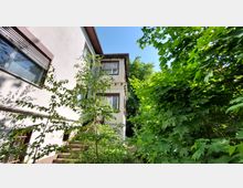 Zweistöckiges Gebäude mit weißen Wänden und braunen Fensterrahmen, teilweise verdeckt von üppigen grünen Bäumen und Sträuchern, unter einem klaren blauen Himmel. Auf der linken Seite sind Fensterläden und metallene Fenstergitter zu sehen.