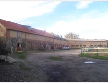 Weitläufiger Hof mit mehreren langgestreckten Gebäuden aus Backstein und Naturstein, teils mit roten Ziegeldächern. Im Vordergrund ein gepflasterter und teils unbefestigter Platz, daneben einzeln stehende Zäune und eine Wiese.