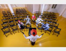 Eine leere Veranstaltungsfläche mit gelbem Boden, auf der viele schwarze Klappstühle in Reihen angeordnet sind. Ein verkleideter Redner mit rotem Helm steht vorne, und einige kostümierte Personen sitzen verteilt auf den Stühlen.