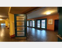 Ein Eingangsbereich mit blau gerahmten Glastüren und teilweise vergitterten Fenstern. Die Wände sind gelb-orange gestrichen, der Boden besteht aus grauen Fliesen, und ein Heizkörper befindet sich neben einer geöffneten Tür.