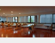 Moderner Seminarraum mit orangefarbenem Boden, langen rechteckigen Tischen und Holzstühlen mit Metallbeinen. Große Fenster mit vertikalen Jalousien lassen Tageslicht herein, und die Decke ist mit runden Einbauleuchten ausgestattet.