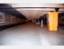 Eine unterirdische Straßenbahnhaltestelle mit Betonboden, Stahlträgern an der Decke und Wänden, die mit Bildern von Straßenbahnen dekoriert sind. Rechts im Bild steht ein orangefarbener Fahrkartenautomat, und im Hintergrund sind Sitzbänke sowie Beleuchtungskörper sichtbar.