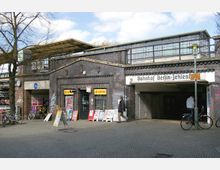 Außenansicht des Bahnhofs Berlin-Zehlendorf mit Backsteinfassade, überdachten Eingängen und Schaufenstern, die unter anderem einen Lottoladen zeigen. Vor dem Gebäude befinden sich eine Pflasterfläche, mehrere Fahrräder und Aufsteller mit Werbung.