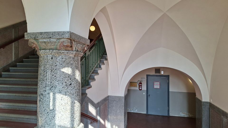 Innenraum eines historischen Treppenhauses mit weißen Kreuzgewölben und rundbogigen Durchgängen, getragen von einer breiten Granitsäule mit verziertem Kapitell. Links führt eine graue Steintreppe mit grünem Geländer nach oben; im Hintergrund befindet sich eine graue Tür in einer gewölbten Nische.