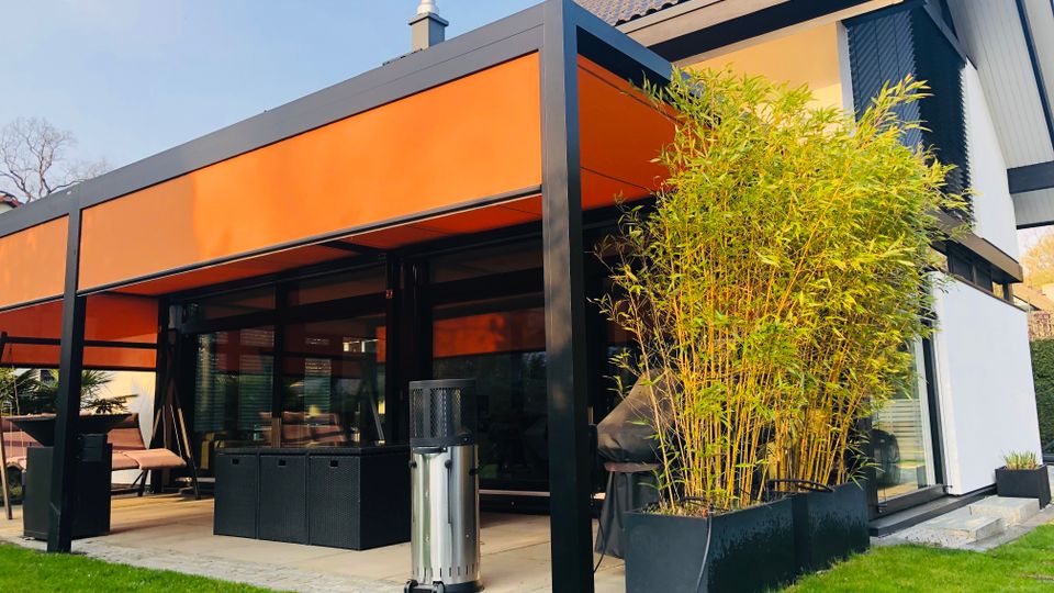 Moderne Hausfassade mit überdachter Terrasse, schwarzem Metallrahmen und orangefarbener Markise vor großen Glasschiebetüren. Daneben stehen ein hoher Bambus im Pflanzkübel, Terrassenmöbel und ein Rasen mit Trittsteinen.