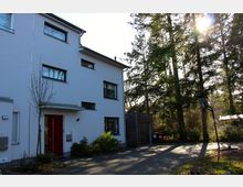 Zweistöckiges, modernes Wohnhaus mit weißer Fassade, roter Eingangstür und mehreren Fenstern. Rechts neben dem Gebäude befinden sich eine Garage und eine Einfahrt, umgeben von hohen Bäumen in einer bewaldeten Umgebung.