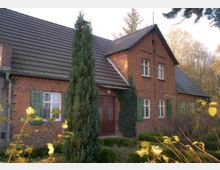 Backsteinhaus mit Satteldach und grünen Fensterläden in einer ländlichen Umgebung. Vor dem Gebäude stehen eine hohe Konifere und Hecken, mit vereinzeltem Efeubewuchs an der Fassade.