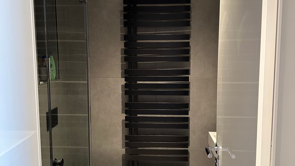 Modernes, grau gefliestes Badezimmer mit einer Glasdusche links und einem wandhängenden WC. An der Rückwand befindet sich ein großer, schwarzer Handtuchheizkörper mit horizontalen Streben.