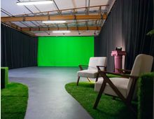 Ein Aufnahmestudio mit grünem Chroma-Key-Hintergrund, umgeben von schwarzen Vorhängen. Im Vordergrund befinden sich zwei gepolsterte Stühle auf einem mit künstlichem Gras bedeckten Boden und eine rosafarbene dekorative Säule.