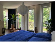 Modernes Schlafzimmer mit großem Bett und blauer Bettwäsche, einer runden Papier-Hängeleuchte sowie Holzboden. Bodentiefe Fenster mit weißen Vorhängen öffnen den Blick auf einen Balkon mit Geländer und viel grünem Baumbestand; mehrere große Zimmerpflanzen stehen am Fenster.