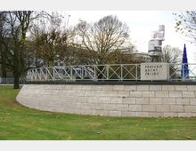 Ein Denkmal mit der Inschrift „Freiheit, Recht, Frieden“, umgeben von einer Metallgeländerstruktur, auf einer erhöhten Plattform aus hellen Steinen; im Hintergrund kahle Bäume und moderne Gebäude mit leuchtenden Elementen.