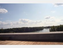Holzterrasse mit niedrigem Steinbrüstungszaun im Vordergrund, dahinter ein Fluss, der von Bäumen gesäumt ist. Am linken Bildrand ist ein Riesenrad im Hintergrund zu erkennen, bei sonnigem Himmel mit vereinzelten Wolken.