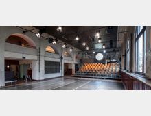 Theaterraum mit einer kleinen, ansteigenden Zuschauertribüne aus Holzstühlen, einem schwarzen Bühnenboden und hellem, leicht abgenutztem Mauerwerk. Die Decke ist mit Beleuchtungskörpern und Kabelinstallationen ausgestattet, während große Fenster auf der rechten Seite Tageslicht einlassen.